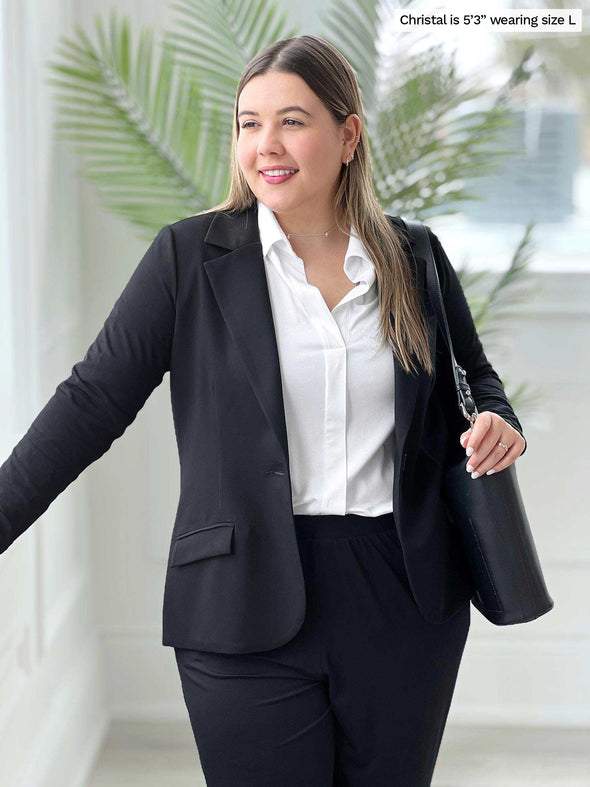 Miik model Christal (5'3", large) leaning against a window looking away wearing Miik's SIenna girlfriend blazer in black with a collared shirt in white and black pants 