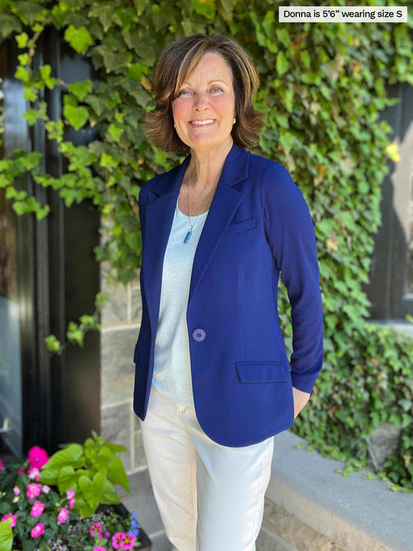 Woman standing in front of nature smiling while wearing Miik's Sienna girlfriend blazer in ink blue colour on top of a light blue top and white pants.