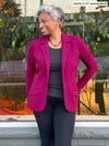 Woman leaning against a window looking away smiling while wearing Miik's Sienna girlfriend blazer in pink colour on top of a grey top with black jeans.