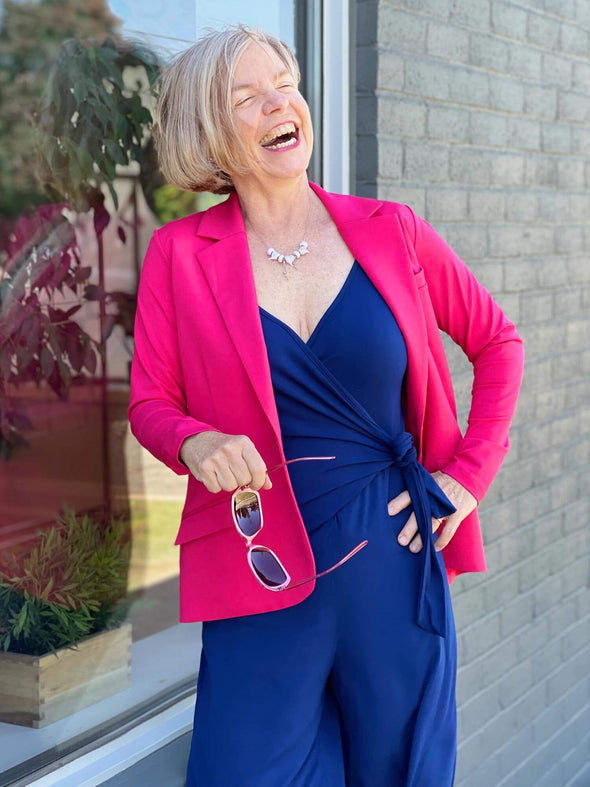 Woman leaning against a window laughing while wearing Miik's Sienna girlfriend blazer in pink raspberry colour with a blue jumpsuit.