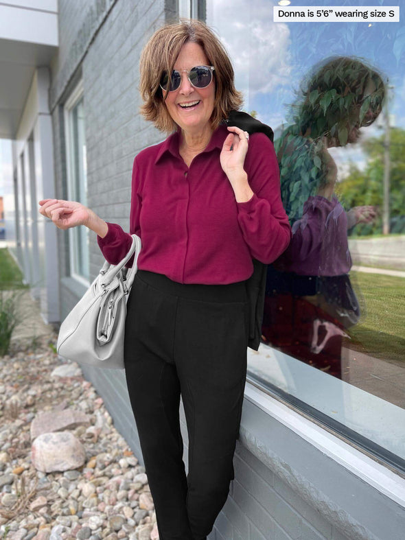 Woman smiling standing next to a window wearing Miik's Silvie slouch pant in black with a collared top in red while holding a purse and a blazer over her shoulders