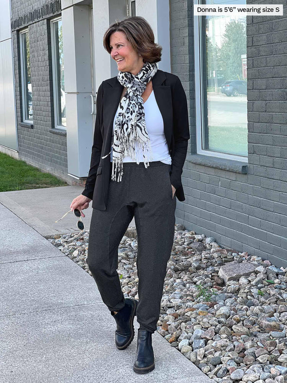 Woman smiling and looking away while standing in a sidewalk wearing Miik's Silvie slouch pant in charcoal with a white top, black blazer and an animal print scarf