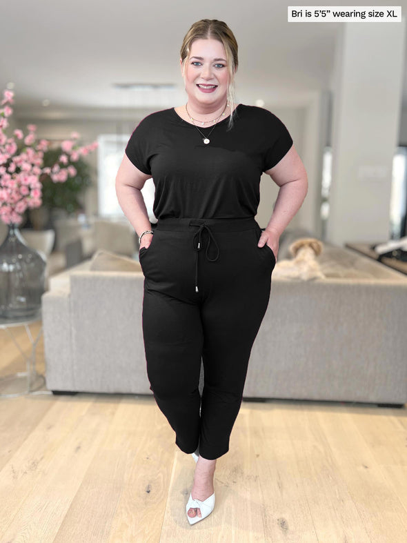 Woman smiling standing in a living room wearing Miik's Stef v-neck open-back capri jumpsuit in black with hands on the pockets 