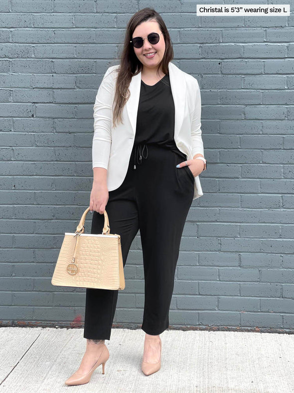 Woman standing in front of a brick wall smiling while wearing Miik's Stef v-neck open-back capri jumpsuit in black with a white blazer on top.