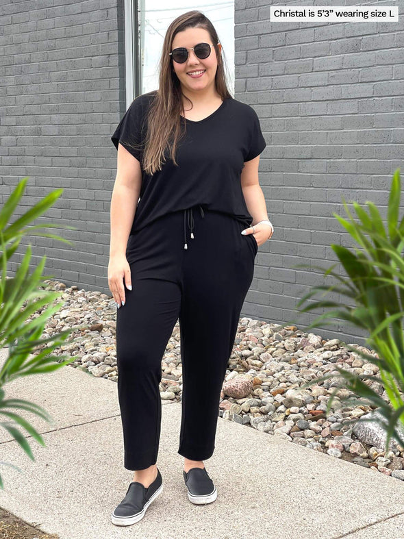 Woman standing outside next to a brick wall smiling while wearing Miik's Stef v-neck open-back capri jumpsuit in black.  