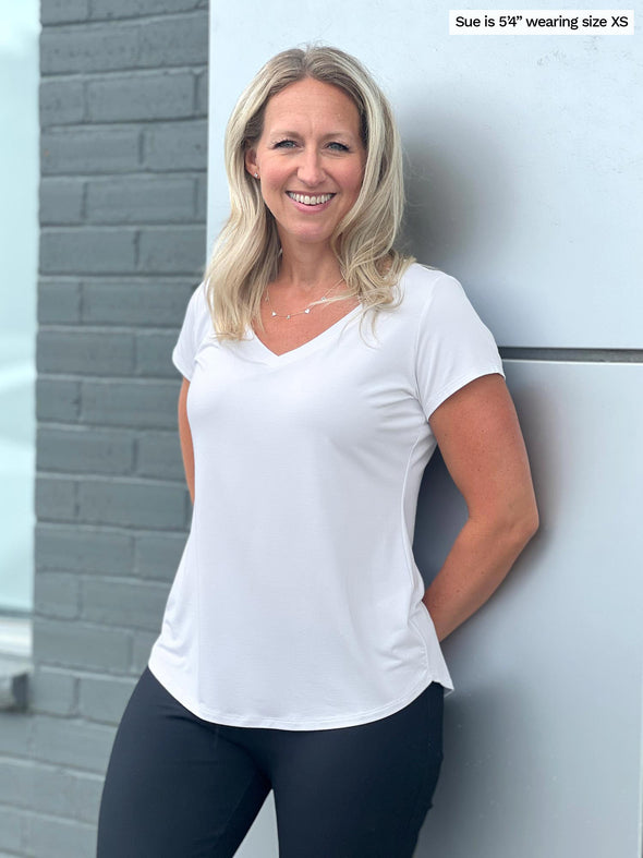 Woman leaning against a grey wall, smiling while wearing Miik's Sutton v-neck classic tee in white with black pants.