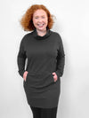 Woman smiling standing in front of a white wall wearing Miik's Tate cowl neck lounge pocket tunic in charcoal with hands on the pockets 
