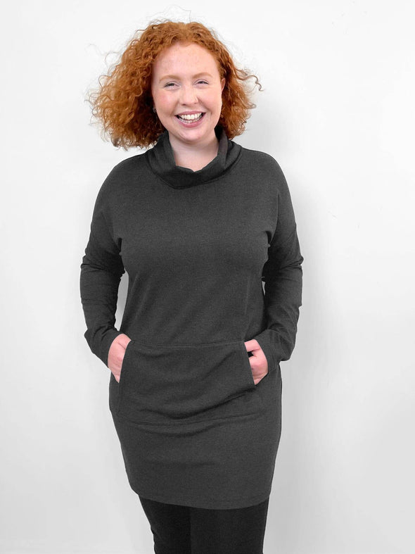 Woman smiling standing in front of a white wall wearing Miik's Tate cowl neck lounge pocket tunic in charcoal with hands on the pockets 