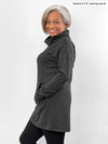 Woman standing sideway in front of a white wall wearing Miik's Tate cowl neck lounge pocket tunic in charcoal with black legging