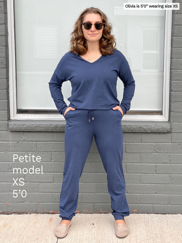 Woman standing in front of a brick wall wearing Miik's Tula long sleeve open-back jumpsuit in navy melange to show how the open back jumpsuit is fitting on a petite model 5”0.