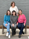 Group of women wearing all the different colours of Miik's Tully reversible fleece dolman sweater: oatmeal, teal melange, granite and pomegranate melange. Three of them are sitting on a bench