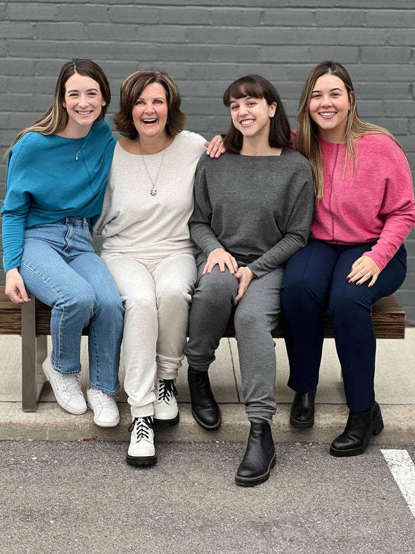 Four women sitting in a bench all wearing all the different colours of Miik's Tully reversible fleece dolman sweater: oatmeal, teal melange, granite and pomegranate melange.