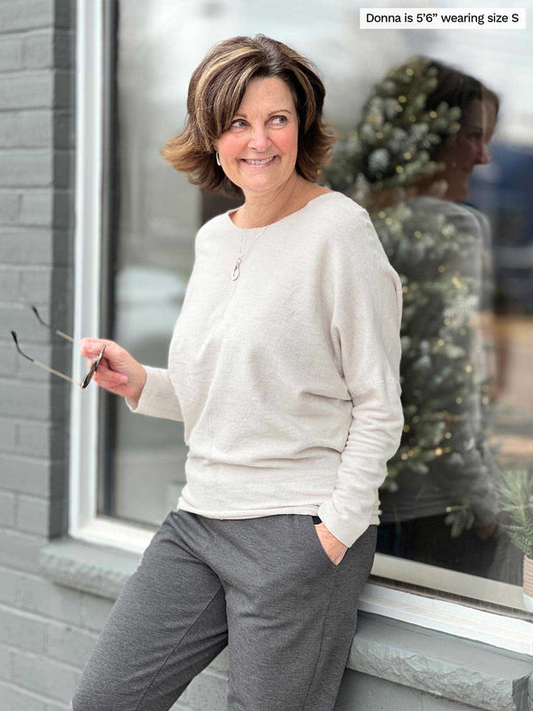 Woman leaning against to a window while looking away wearing  Miik's Tully reversible fleece dolman sweater in oatmeal melange and charcoal pants 