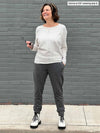 Woman standing in front of a brick wall wearing  Miik's Tully reversible fleece dolman sweater in oatmeal melange with a charcoal pant and boots 