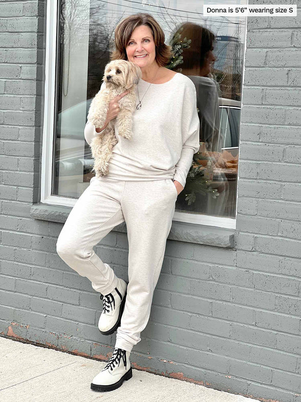 Woman leaning against to a window while holding a puppy wearing  Miik's Tully reversible fleece dolman sweater in oatmeal melange with a matching colour fleece jogger