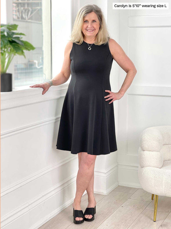 Miik model Carolyn (five feet ten, size large) smiling standing next to a window wearing Miik's Valerie sleeveless fit and flare dress in graphite