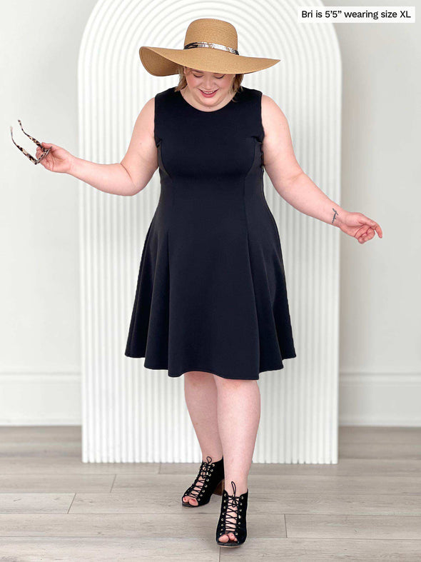 Miik model Bri (five feet five, size extra large) smiling and looking down wearing Miik's Valerie sleeveless fit and flare dress in graphite with a summer hat and black heels 