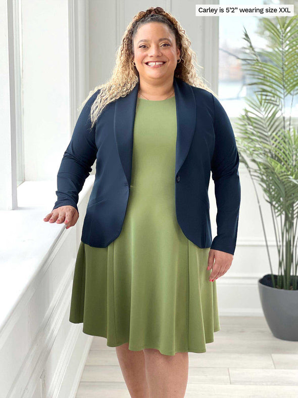 Miik model Carley (five feet two, size double extra large) smiling standing next to a window wearing Miik's Valerie sleeveless fit and flare dress in green moss along with a navy blazer opened