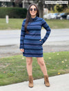 Woman smiling while standing in a sidewalk wearing Miik's Venice cowl pocket tunic in navy jewel tone stripe with boots and sunglasses