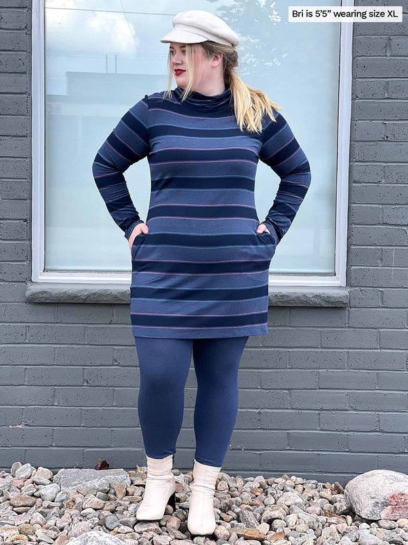 Woman standing in front of a window wearing Miik's Venice cowl pocket tunic in navy jewel tone stripe and navy melange legging