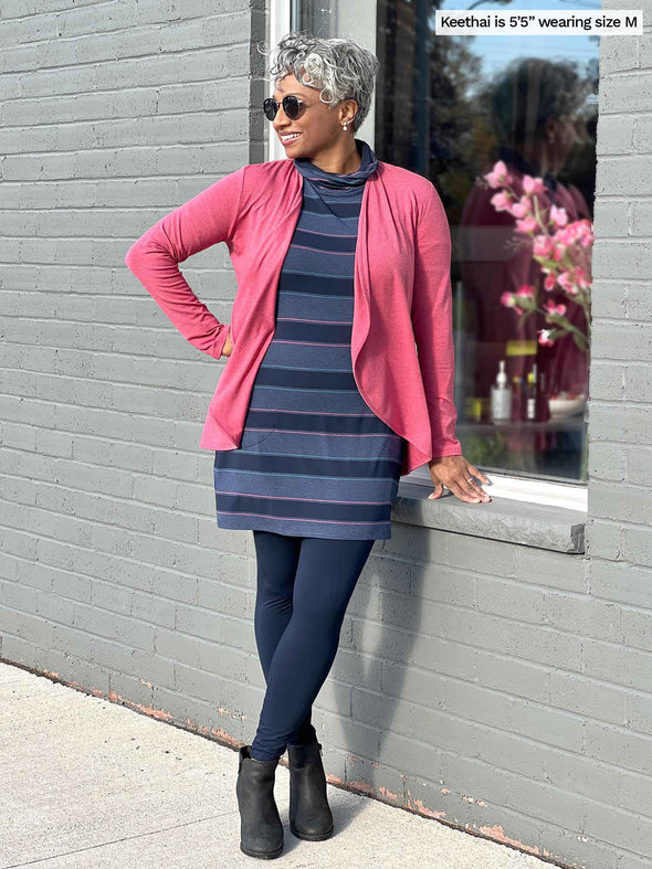 Woman standing in front of a window wearing Miik's Venice cowl pocket tunic in navy jewel tone stripe, navy legging and a pink pomegranate cardigan 