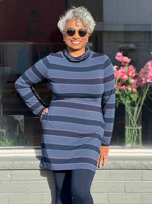 Woman standing in front of a window wearing Miik's Venice cowl pocket tunic in navy jewel tone stripe, navy legging and sunglasses 