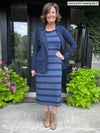 Woman standing in a doorway while wearing Miik's Verona long sleeve boat neck dress in navy jewel tone stripe with a blazer in navy melange and boots