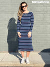 Miik model Johanna (five foot six, xsmall) smiling while standing in front of a brick wall wearing Miik's Verona long sleeve boat neck dress in navy jewel stripe and sneakers 