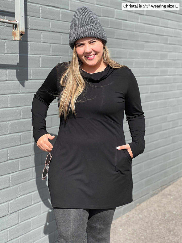 Woman standing in front of a brick wall wearing Miik's Vienna cowl pocket tunic in black with charcoal legging and toque