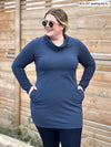 Woman standing in front of a wood wall looking away wearing Miik's Vienna cowl pocket tunic in navy melange with a legging in navy and sunglasses