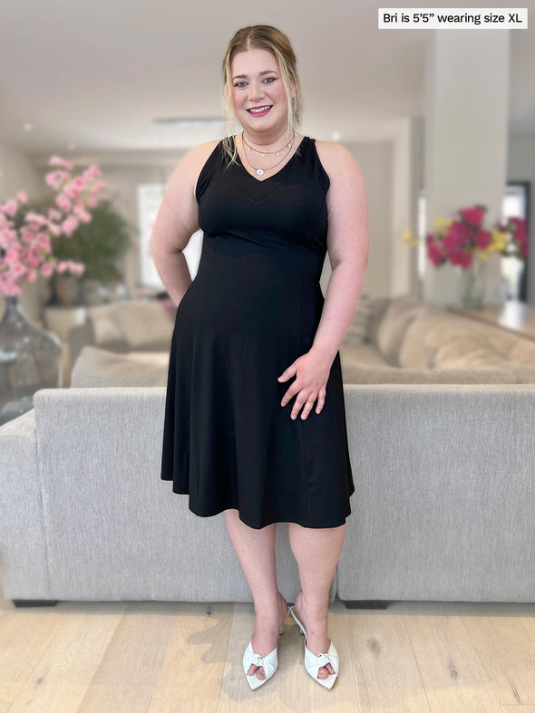 Woman smiling standing in a living room wearing Miik's Zayd v-neck racerback dress in black with white heels and accessories 