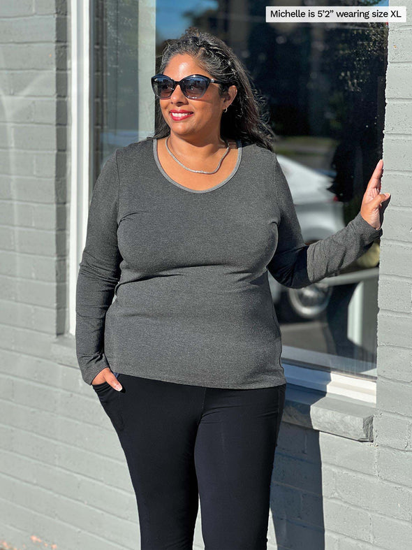 Woman standing outside leaning against the window of a brick building, smiling while wearing shades and Miik's Albany reverisble long sleeve tee in charcoal with black pants.