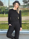 Woman standing in front of a window wearing an all black outfit: Miik's Sigourney asymmetrical drape neck top and a jogger 