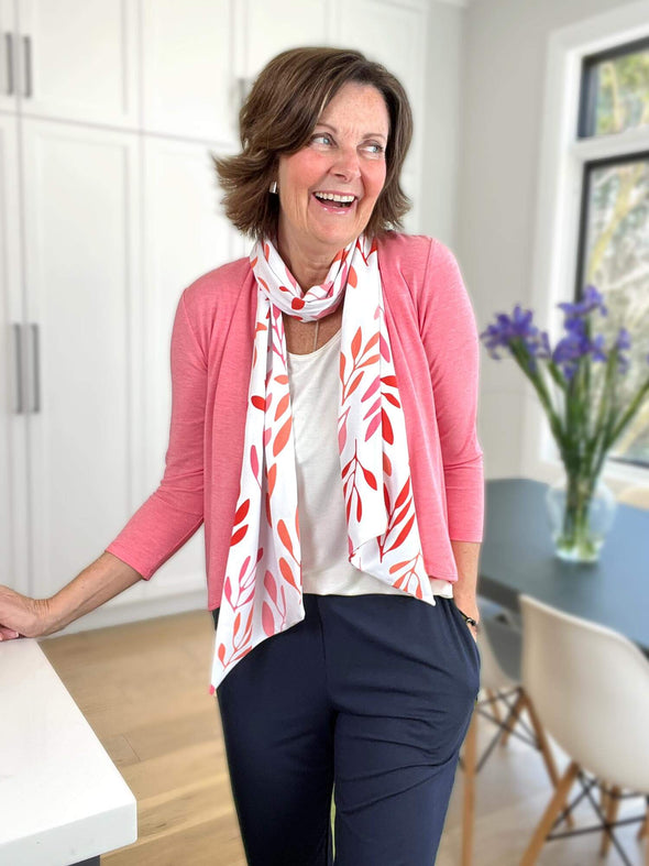 Woman standing next to a dining table looking away smiling wearing a white top with a pink cardigan overtop, pairing it with Miik's Halona tie scarf with pink leaf pattern and navy pants.