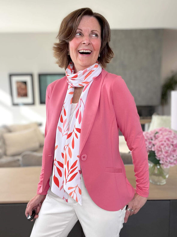 Woman standing in the leaving room looking away laughing wearing a white top and pants, with a pinkk blazer on top and Miik's Halona tie scarf in pink leaf pattern tied around her neck.