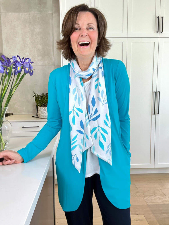 Woman standing in the kitchen laughing wearing a white top with Miik's Halona tie scarf in blue leaf pattern around her neck and a matching colour cardigan.