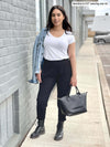 Woman standing on a sidewalk against a grey brick building while wearing Miik's Silvie slouch pant in black with a white tee and jean jacket.